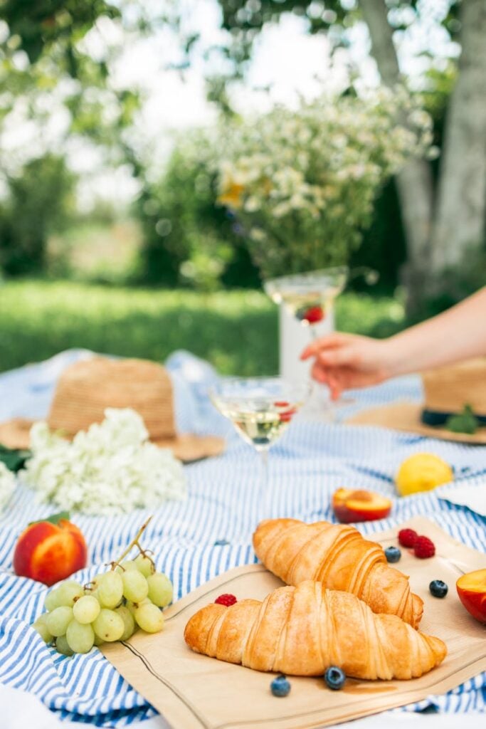 person holding a glass of white wine beside fruits and bread on table